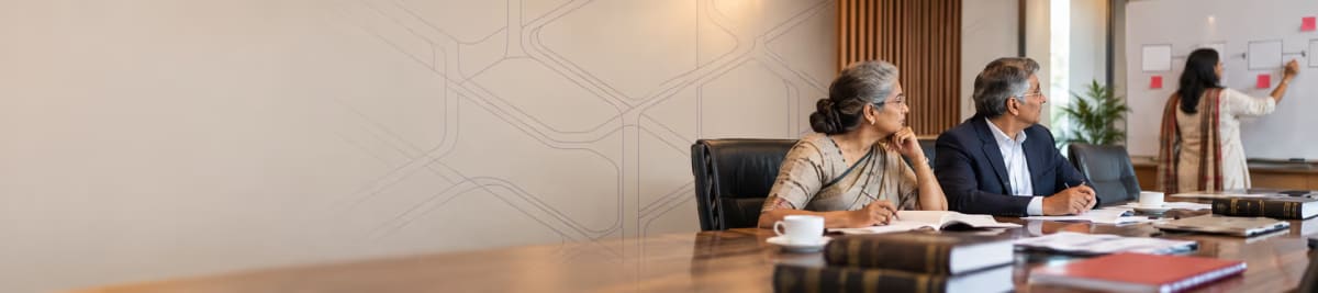 Two senior colleagues seated at a boardroom table with open files and notebooks, watching a third person map out a plan on a whiteboard behind them