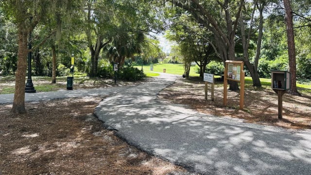 The paved trail at Arlington Park curving past a trail information kiosk and a Little Free Library, under moss-draped oaks