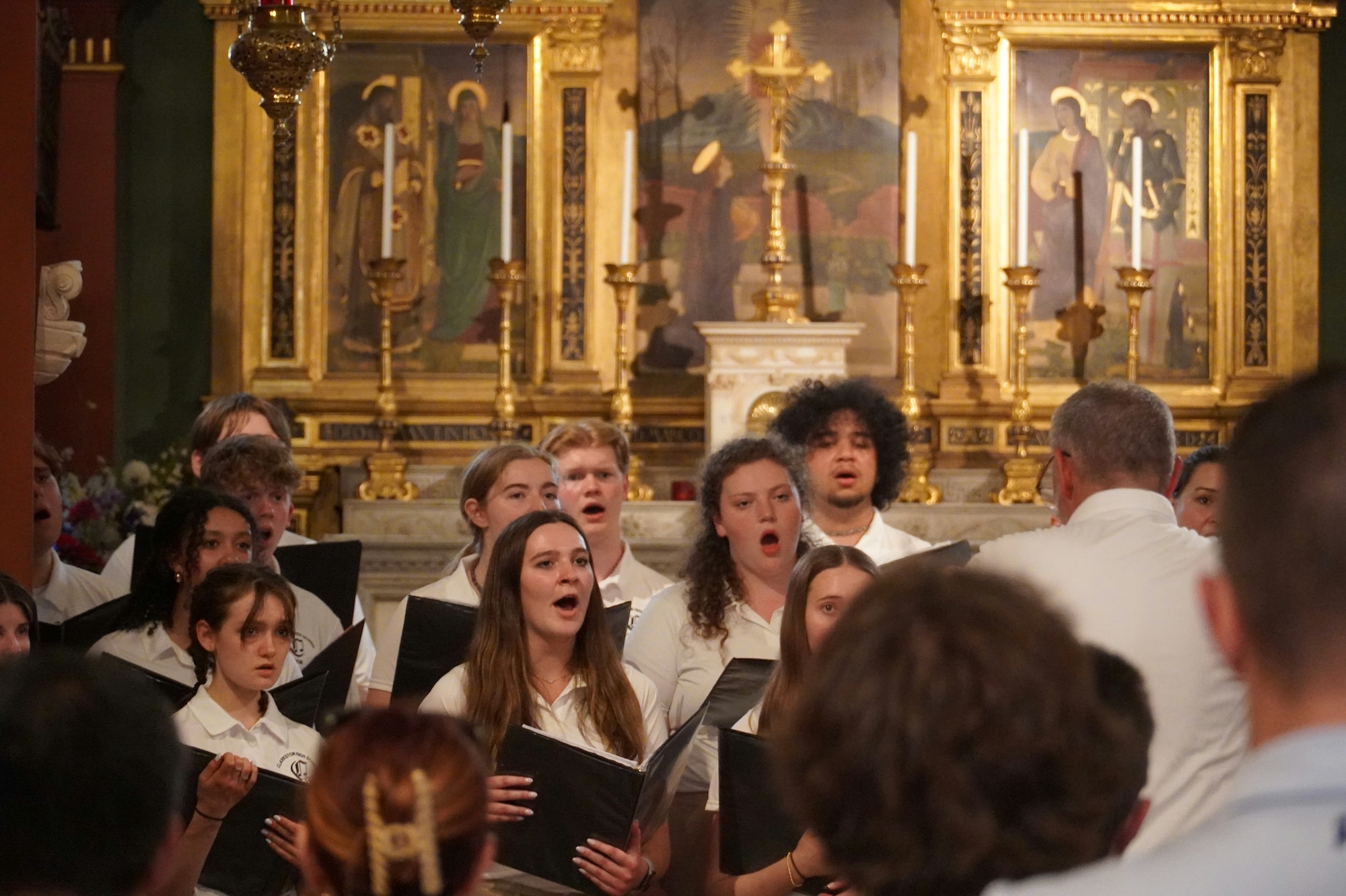 A college choir performs in a church, with an ornate altar in the background.