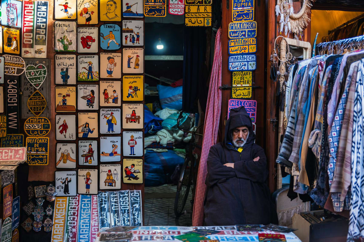 A vendor in Marrakech souk, surrounded by vibrant signs and garments, embodies local culture.