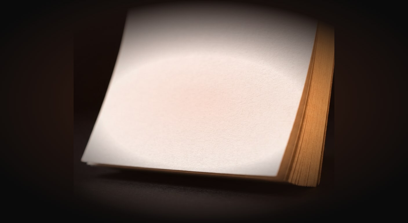 Macro close-up of a single tear-off desk calendar page standing upright on a near-black surface, very shallow depth of field with crisp paper texture and a softly curling corner, one clean blank page centered with generous empty dark margins all around, warm amber rim light catch