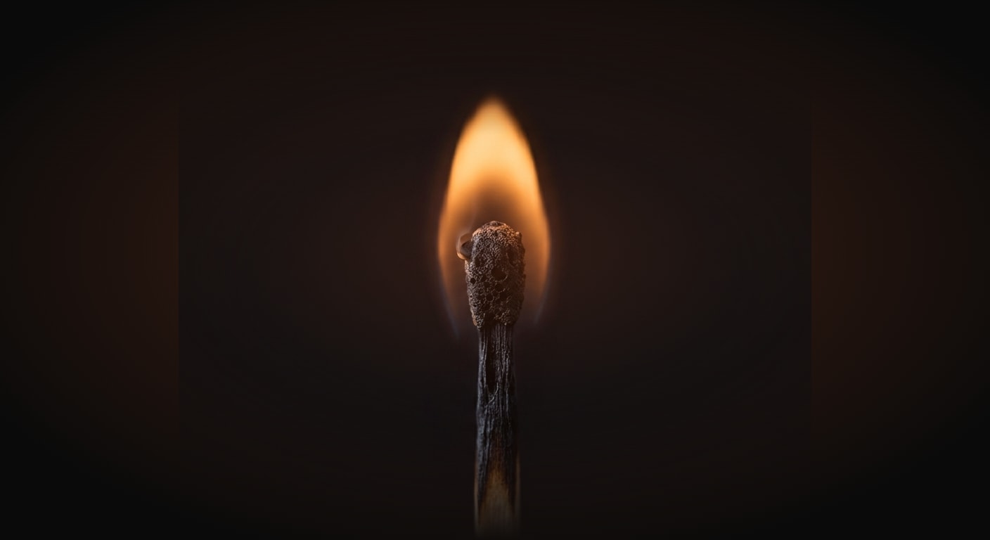Macro close-up product shot of a single burning matchstick standing upright, its flame glowing warm amber against a near-black background (#0A0A0A), very shallow depth of field with crisp texture on the charred wood and bright flame, symbolizing how easily a streak burns out.