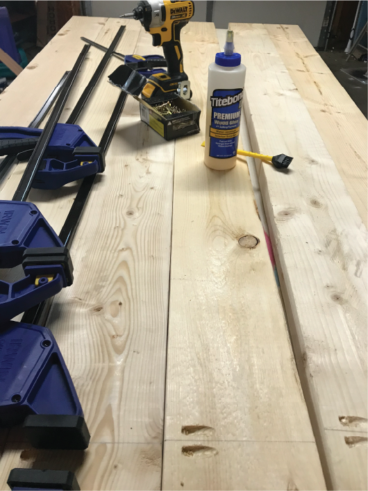 Boards laid on a table with clamps, wood glue and a drill sitting on them.