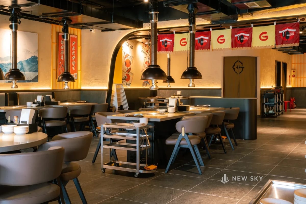 Dải cờ vải đỏ và vàng treo dưới trần sơn đen còn hở nguyên đường ống, phía dưới là dãy bàn nướng có ống hút khói chạy thẳng lên.