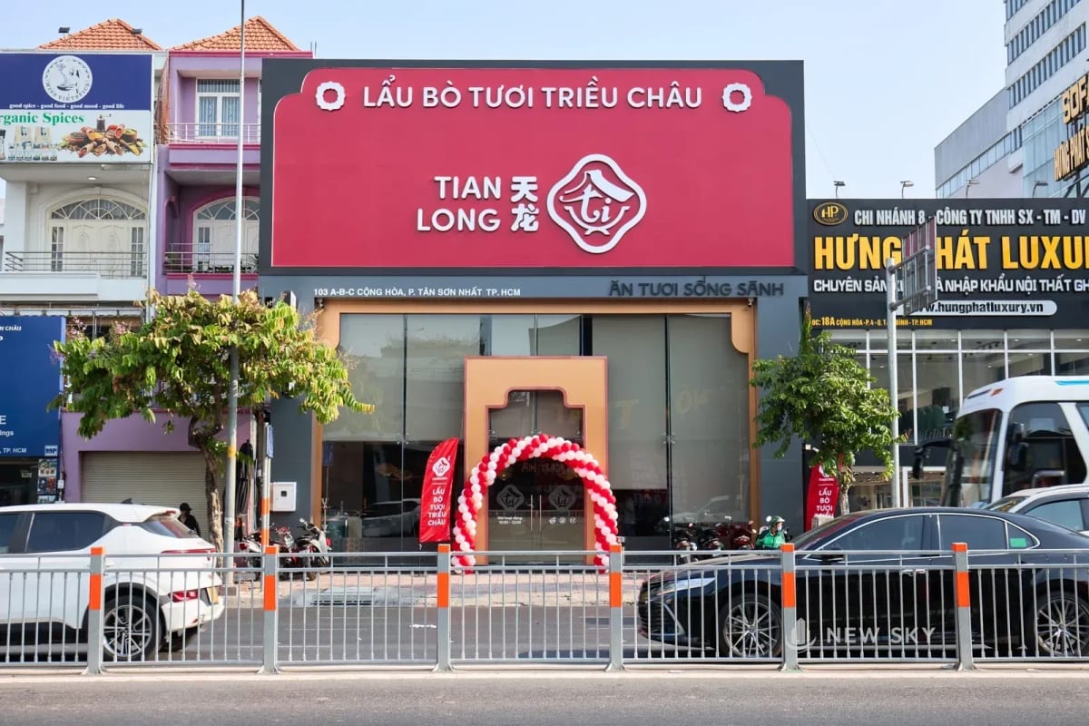 Mặt trước nhà hàng Tian Long trên đường Cộng Hòa: tấm biển đỏ khổ lớn nằm ngang trên đầu tầng trệt, dưới là mặt kính chạy suốt và cửa vào đóng khung bằng một vòm màu đồng; ngoài đường có hàng rào sắt phân làn.