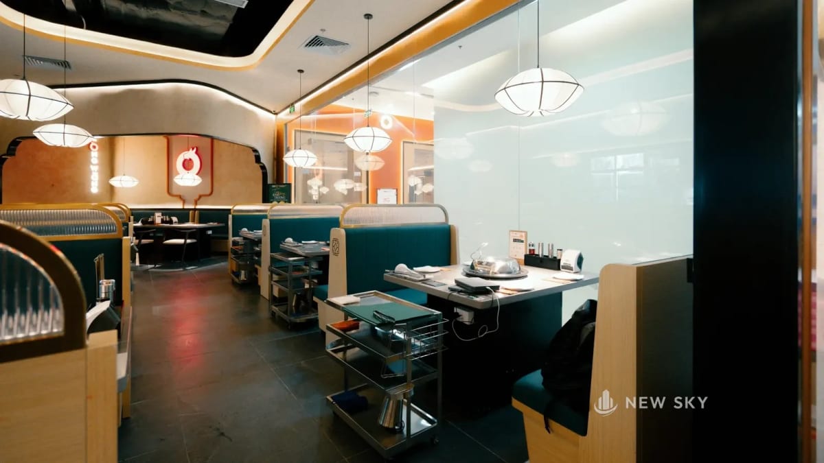 Phòng ăn Long Wang Vincom 191 Bà Triệu nhìn dọc chiều dài: các khoang ghế băng xanh đậm xếp nối nhau sát vách kính, trần viền thạch cao màu kem, cuối phòng là khoang bàn sau một khung vòm, logo tròn đỏ trên tường cam.