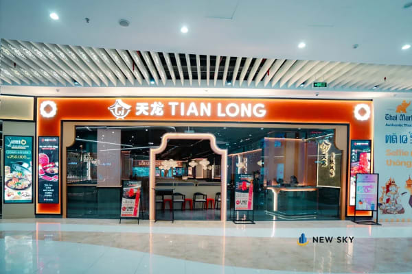 Lối vào gian Tian Long nhìn từ hành lang trung tâm thương mại: một vòm lớn đóng khung cửa, mảng tường quanh vòm màu cam đỏ, tên thương hiệu nằm trên đỉnh vòm; trần hành lang là nan trắng.