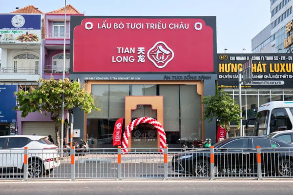 Mặt trước nhà hàng Tian Long trên đường Cộng Hòa: tấm biển đỏ khổ lớn nằm ngang trên đầu tầng trệt, dưới là mặt kính chạy suốt và cửa vào đóng khung bằng một vòm màu đồng; ngoài đường có hàng rào sắt phân làn.