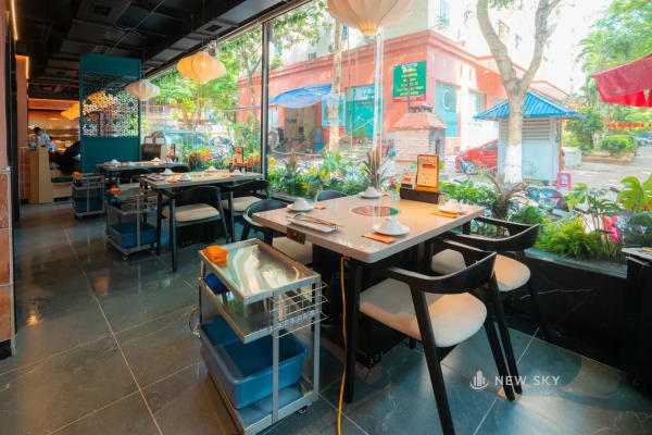 Bàn ăn kê sát mặt kính, giữa bàn là ô bếp tròn; ngay bên trong kính là một bồn cây trồng cây lá to cao ngang mặt bàn, ngoài kính là hàng cây và nhà cửa bên đường.