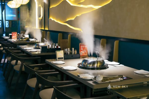 Ba cột hơi trắng bốc lên từ các nồi hấp giữa bàn, phía sau là mảng tường phủ ánh vàng đồng có nét sáng hình dãy núi chạy ngang; bên phải là dãy ghế băng, bên trái là ghế rời.