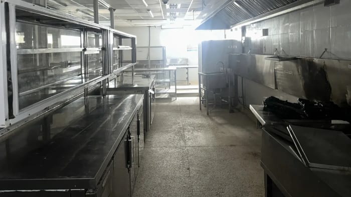 Gian bếp dài của một mặt bằng cũ: bên trái là dãy bàn inox có cánh tủ bên dưới, phía trên là giá khung inox lắp kính; bên phải là chụp hút khói inox chạy dọc tường, một chậu rửa đứng chân cao và chỗ đặt bếp nấu, tường phía sau ám khói đen; cuối gian có thêm bàn và kệ inox, ánh sáng hắt vào từ cửa sổ.