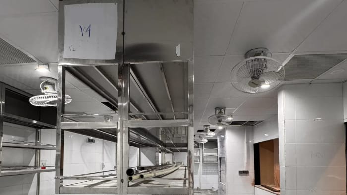Kệ inox treo dưới trần trong một khu bếp đang thi công, tường ốp gạch men trắng, quạt treo trần; trên thân kệ dán tờ giấy viết tay mã V4.