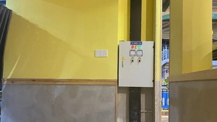 Một tủ điện vỏ trắng gắn trên tường sơn vàng, nhãn nền xanh đậm ghi "TỦ ĐIỆN TD2", mặt tủ có ba đèn báo đỏ, vàng, xanh và hai đồng hồ đo. Bên trái tủ là ba công tắc đèn; chân tường bên dưới tô xám, ngăn với phần sơn vàng bằng một nẹp gỗ chạy ngang.