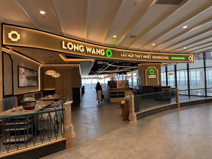 Mặt tiền nhà hàng Long Wang ở tầng 4 Aeon Mall Thanh Hóa: dải biển cong chữ LONG WANG với logo tròn xanh lá và dòng Lẩu hấp thuỷ nhiệt Hongkong, lan can song hoa văn xanh đậm, bên trong là tranh thủy mặc, đèn lồng trắng và vách kính nhìn ra ngoài trời; bàn ghế còn bọc ni lông.