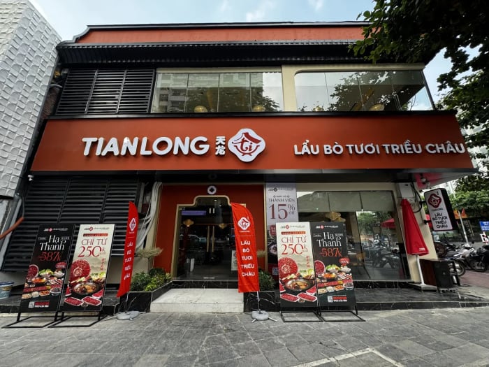 Mặt tiền nhà hàng Tian Long số 10 Huỳnh Thúc Kháng: dải biển nền đỏ cam chữ TIANLONG và LẨU BÒ TƯƠI TRIỀU CHÂU, cửa chính bằng kính, khung viền vàng đỉnh lượn bậc, hai bên là các tấm standee món lẩu và cờ đỏ, tầng 2 là vách kính dài, mép trái là mảng lam đen.