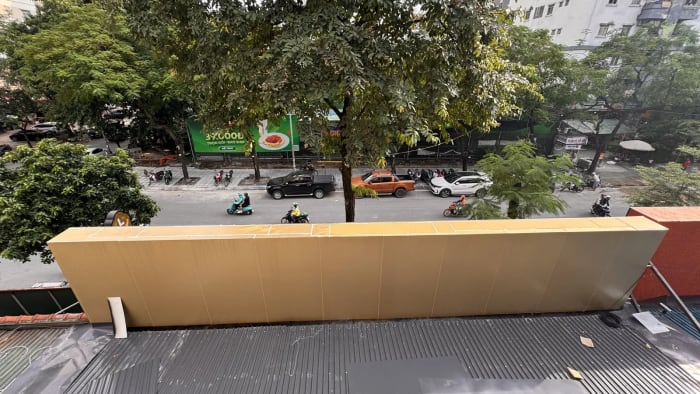 Nhìn từ mái tôn xuống lòng đường: mặt sau phần đỉnh mặt tiền ốp tấm alu màu vàng đồng chạy dài ngang khung, các đường ghép giữa tấm thấy rõ; dưới là mái tôn còn vài tấm che, bên kia đường là hàng cây, ô tô đỗ và tòa chung cư.