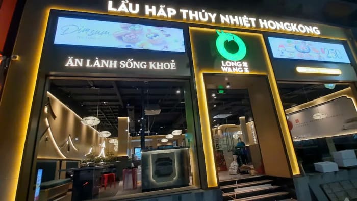 Mặt tiền Long Wang Linh Đàm buổi tối sau khi tháo tấm quây: dải biển sáng chữ LẨU HẤP THỦY NHIỆT HONGKONG, khung viền vàng sáng đèn quanh các ô kính; qua ô kính bên trái thấy ghế băng bọc đệm xanh đậm kê sát kính, sâu trong phòng là đèn lồng trắng thả từ trần sơn tối, tường màu kem có đường mái cong hắt đèn; trước bậc cửa còn vài thùng vật tư.