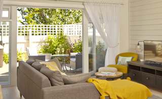 An open-plan view of one of our cottages, with a round table to the right, a brown sofa facing a tv on a unit. Next to the sofa is a set of french doors which are opened up onto a decking area. On the decking is a garden table and chairs next to a hot tub image