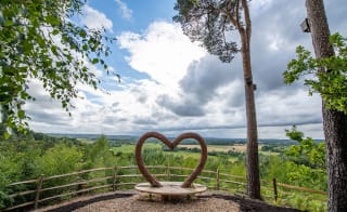 A panoramic view of the countryside, with a viewing area in the foreground. The bench features a wooden heart arch. image