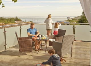 A family of four on the balcony of their holiday home in The Bay Colwell. There are two adults, a young boy, and a young girl with strawberry blonde hair, and all dressed in summer attire. image