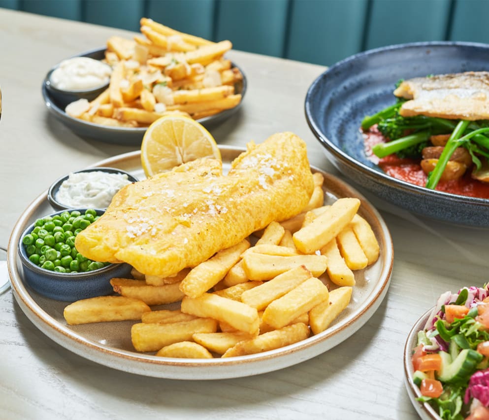 A close up of fish and chips on a wooden table with various other dishes around it.