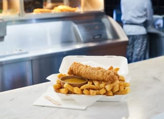 An image of a battered sausage on chips with a pot of curry sauce at Catch 22. In the background there is a chef cooking. image