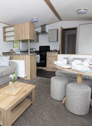 Suggested: The interior of one of our luxury caravans. There's a large, grey sofa, a seating area with a table, and a kitchen space with several appliances.