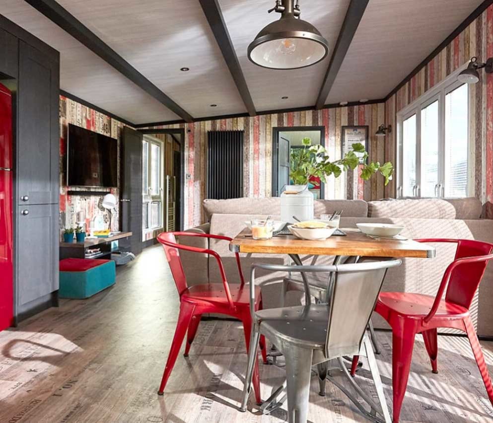 An image of the interior of one of our holiday lodges. There's wooden panneling, a cream fabric sofa and red metal dining chairs around a wooden table.