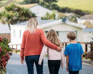 A woman with blonde hair wearing an orange jumper and jeans has her arm around a girl with light brown hair, who is wearing a pale pink top and black trousers. They are walking together down a path at our holiday park, Newquay Bay, Cornwall. image