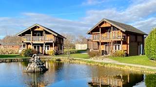 Two of our stunning lodges at Woodland Lakes, overlooking onto a small lake on a sunny, cloudy day with blue skies. image
