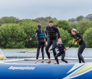 A young family wearing life jackets and full wetsuits balancing on a inflateable in a body of water at one of our holiday parks.