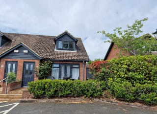 An outside view of one of our 3-bed cottages. Bushes outside lead to three steps to the pathway of the front door to the cottage. The sky above is blue yet very cloudy. image