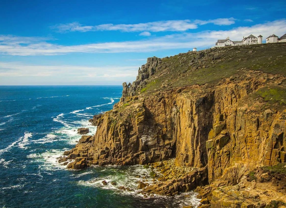 A landscape image of the sea and cliffs located at Lands End. A blue sky with minimal clouds sets the scene with the Land's end tourist building seen in the top right of the image. image