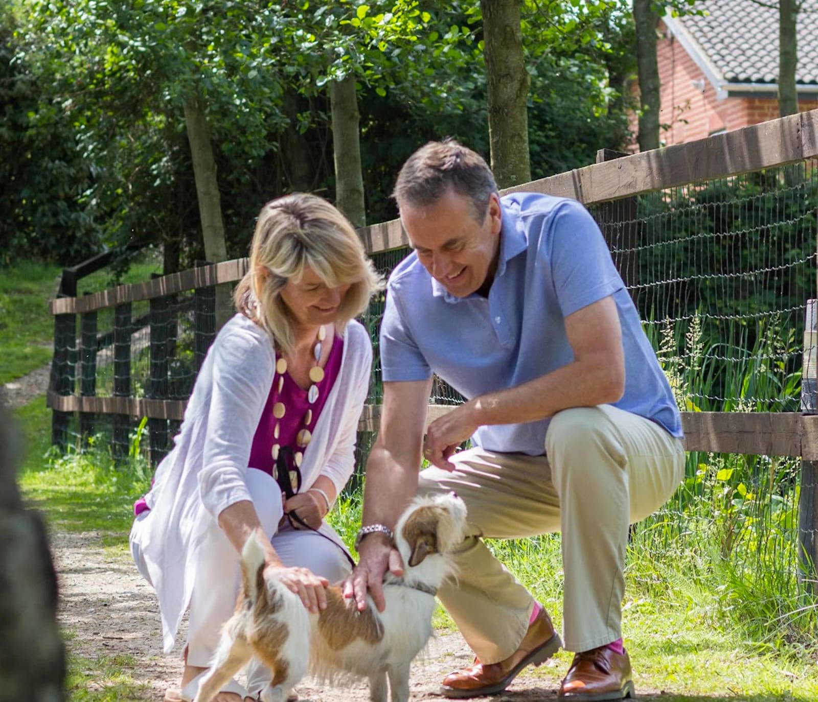 A mature man and woman are petting a light brown and white fluffy dog. Both the man and woman are kneeling down and smiling.