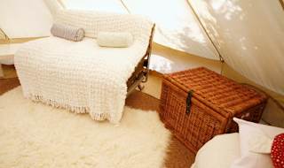 A photo of the living space inside of a canvas bell tent. To the middle and right side of the photo is two chairs, one has a thick white cover with two white cylinder cushions and the other has a white square pillow resting on the arm of the c image