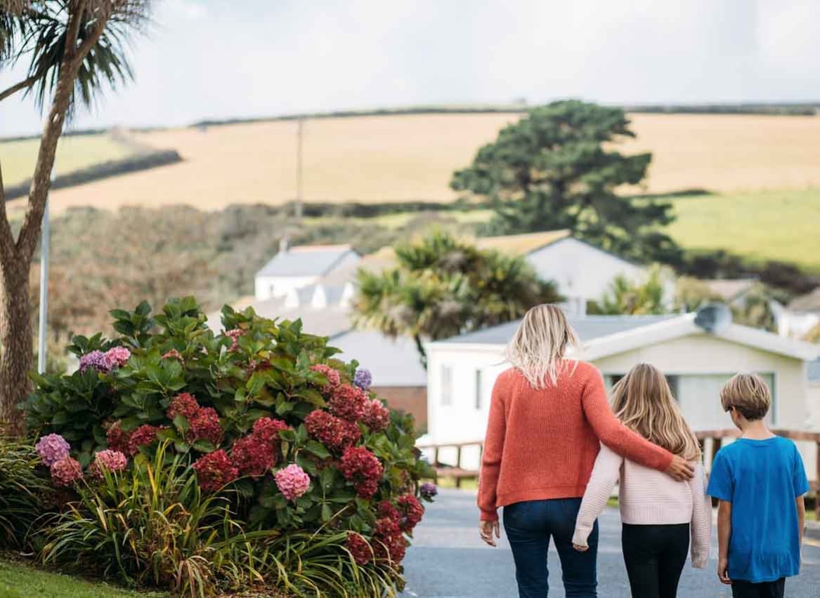 A woman with blonde hair wearing an orange jumper and jeans has her arm around a girl with light brown hair, who is wearing a pale pink top and black trousers. They are walking together down a path at our holiday park, Newquay Bay, Cornwall. image