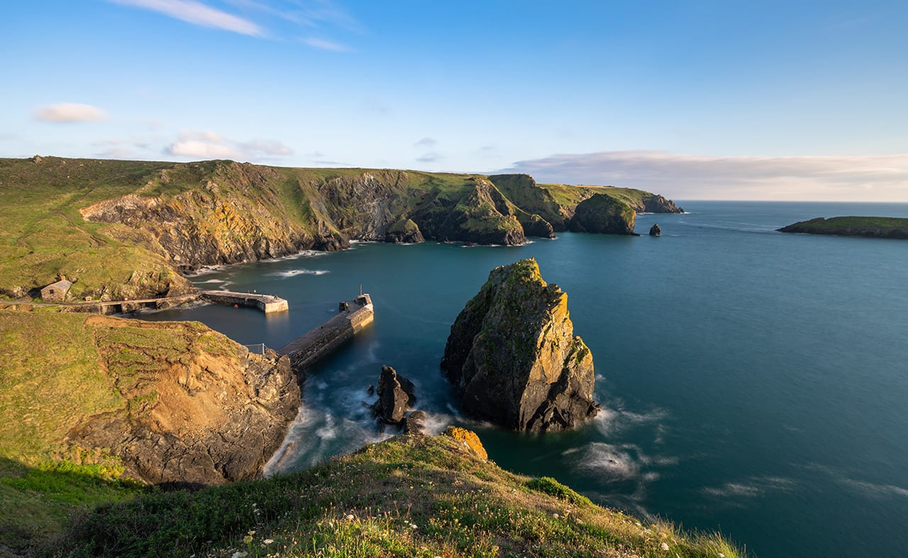 An image of Mullion Cove, Cornwall on a sunny  day. There are views of the vast cliffs, rocks and stunning blue sea. image