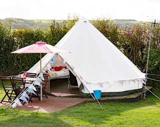 An off white canvas bell tent pitched up outside on the grass on a sunny day. The entrance to the bell tent is open and there is a pink parasol resting against the entrance.