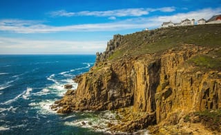 A landscape image of the sea and cliffs located at Lands End. A blue sky with minimal clouds sets the scene with the Land's end tourist building seen in the top right of the image. image
