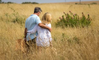 A couple walking in a tall grass field, arm in arm, carrying a wicker picnic basket. image