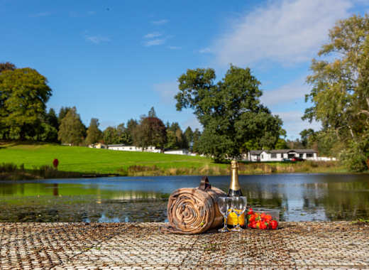 Views over the lake at Moffat Manor Holiday Park. There is a picnic set up with a bottle of prosecco, two glasses, some strawberries and a picnic blanket. There are views of the caravans in the background.