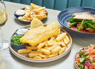 A close up of fish and chips on a wooden table with various other dishes around it. image