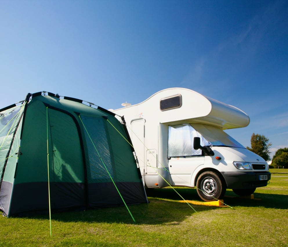 A tent and tourer in one of our holiday parks. The large white tourer is parked next to a green tent, situated on the grass on a sunny day.