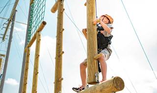 A pre-teen child is climbing on a wodden structure with a bright orange helmet, torso harness and a pully rope system. The child is looking up to judge his next move as he accends higher on the structure.