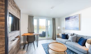 A living and dining area of one of our apartments, contains a three-seater sofa, a round coffee table, a TV mounted into the wall and a small dining table. In the background is a set of french doors leading to a balcony area. image