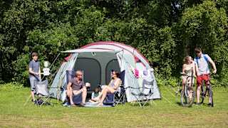 A pitched up family tent with a man, woman and baby sitting in the porch area of the tent. A teen boy and girl are cycling by on a hot sunny day.