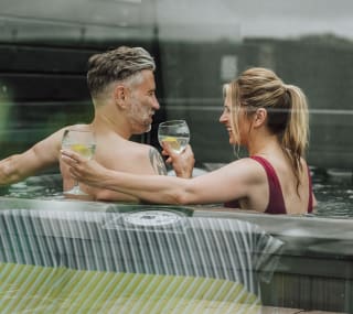 A middle-aged couple are cuddled with drinks in hand, whilst sitting in a hot tub. image