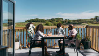 A family sat outside on the decking of their holiday home on a sunny day at The Bay Filey, Yorkshire, overlooking the lake and woodland views.