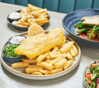 A close up of fish and chips on a wooden table with various other dishes around it. image