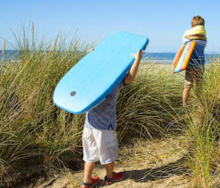 Two children walking across the sand to the sea holiday two body boards on a sunny day.